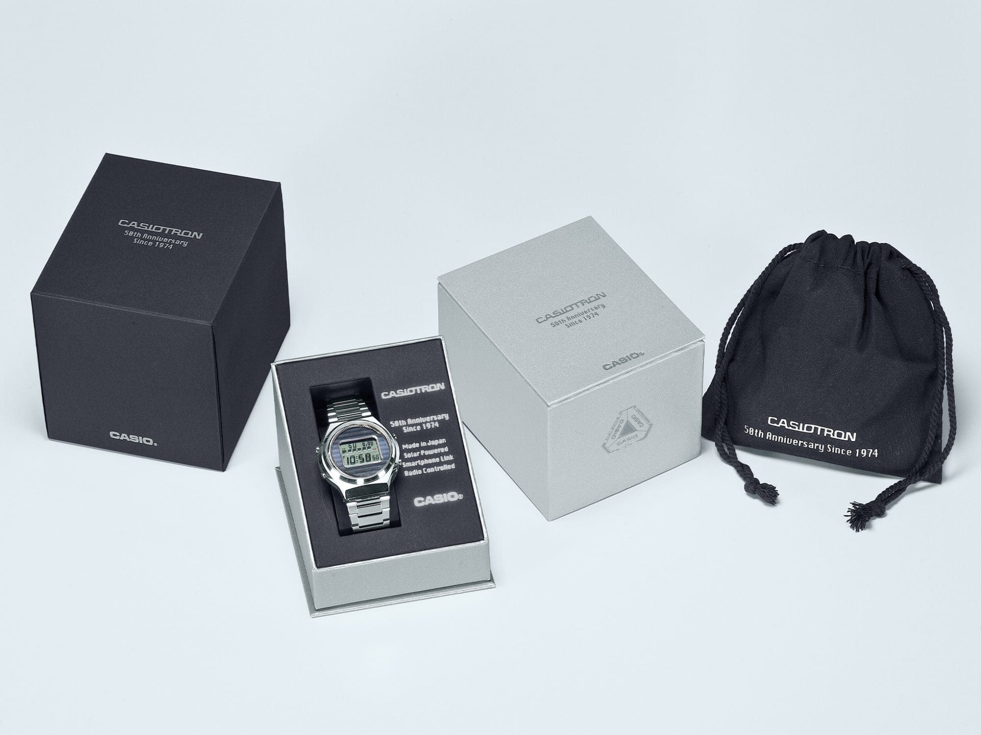 El reloj Casio Casiotron plateado de acero inoxidable en una caja abierta, junto a él la tapa plateada de la caja, el embalaje exterior negro y una bolsa de tela negra. Todas las partes del embalaje están etiquetadas: Casiotron 50 aniversario desde 1974.