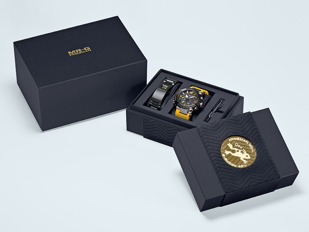 La caja exterior oscura con el logotipo MR-G Frogman en relieve dorado junto a la caja abierta con el reloj MRG-BF1000E-1A9 con correa amarilla. Además, la caja contiene la correa de acero inoxidable y la herramienta de montaje.