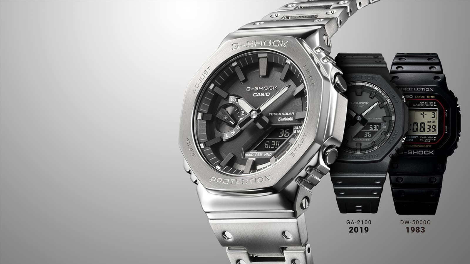 El reloj plateado de acero inoxidable Casio G-Shock GM-B2100D con esfera negra delante del reloj híbrido GA-2100 negro de 2019 y del reloj digital negro DW.5000C de 1983