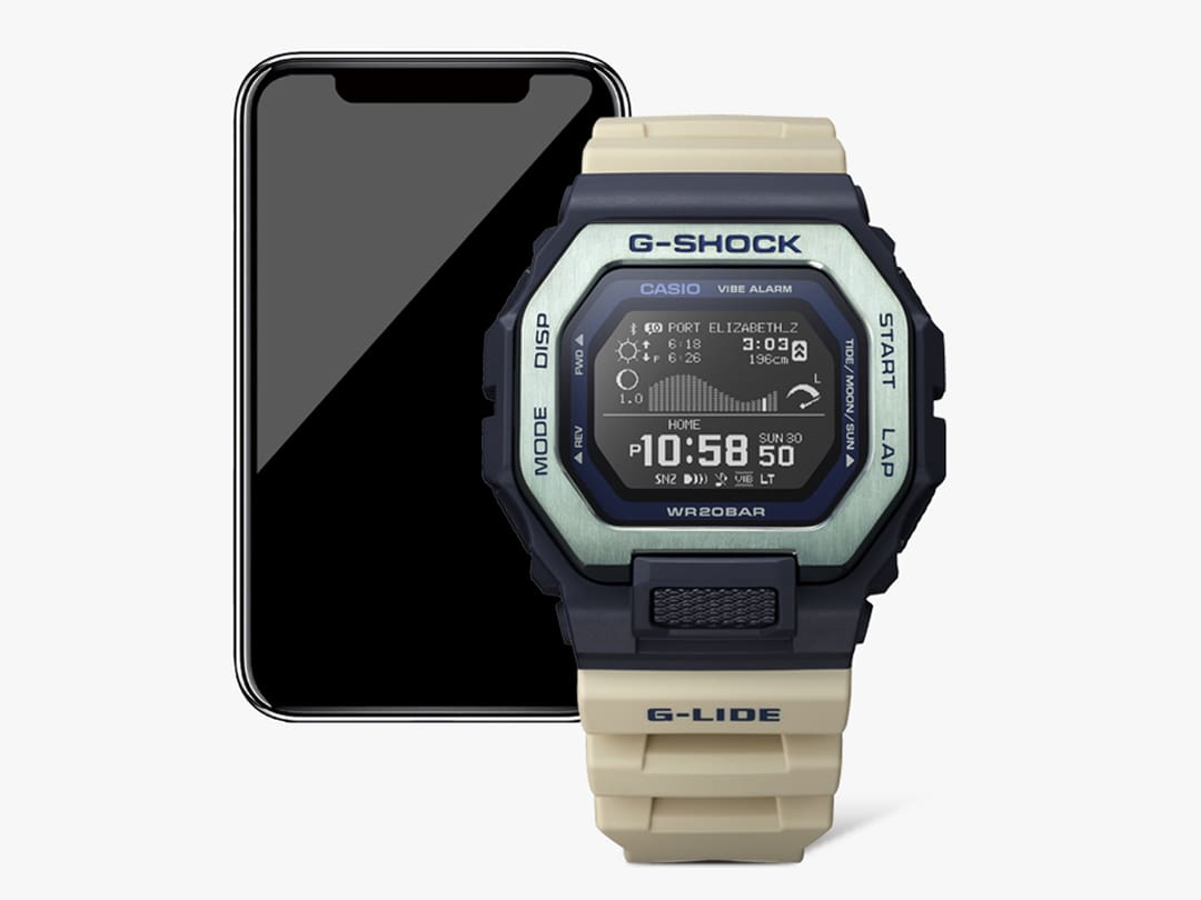 Delante: un reloj Casio GBX-100-2 con correa beige, carcasa negra y bisel de acero inoxidable plateado; detrás: un smartphone muestra información sobre el spot de surf de Port Elizabeth, en Sudáfrica, en la aplicación G-Shock Move.