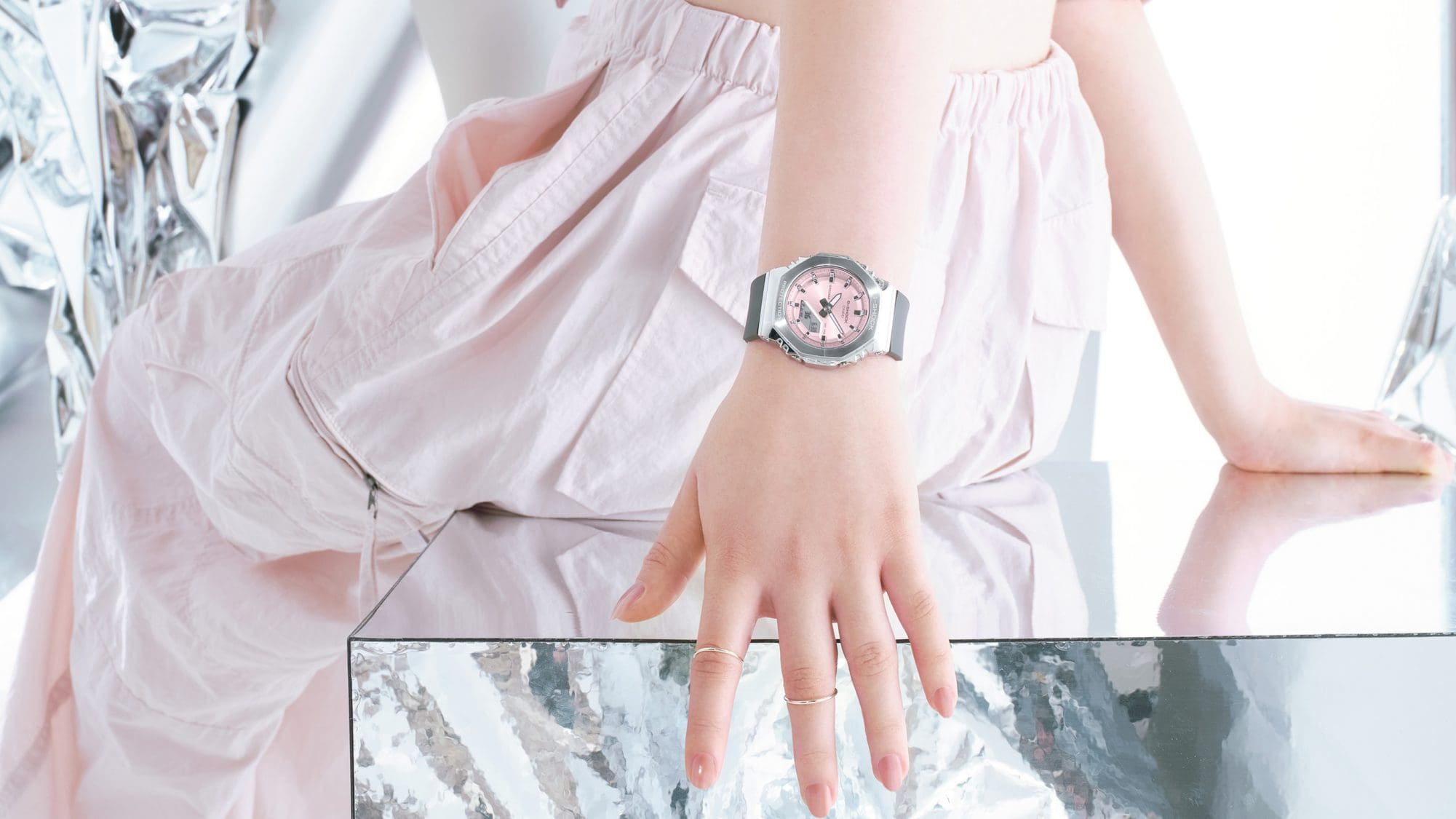El reloj de pulsera Casio G-Shock GM-S2110-4A con esfera rosa, caja plateada y correa negra en la muñeca de una mujer sentada en un taburete de espejo