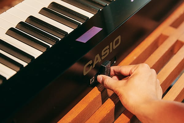 Una mano inserta un adaptador MIDI y audio en el piano digital Privia PX-S3100 negro de CASIO