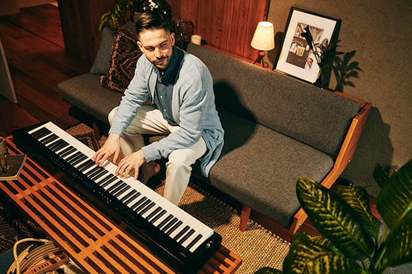 Un joven está sentado en un sofá. Delante de él, sobre la mesa del salón, está el piano digital Privia PX-S3100BK en el que toca el piano