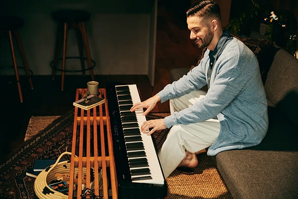 Un joven sonriente sentado en un sofá toca el piano digital Privia PX-S3100BK que tiene delante sobre la mesa del salón