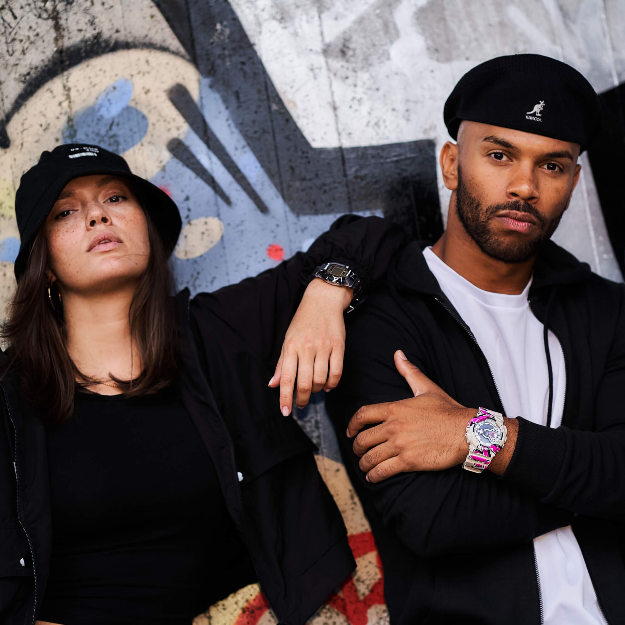 Dos personas se apoyan en un muro de hormigón pintado con spray, con ropa oscura y llamativos relojes G-SHOCK.