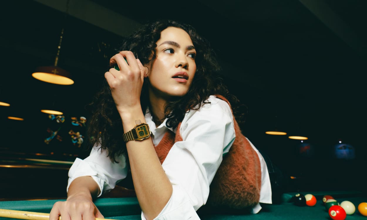 A130WEG-9AEF Une femme vêtue d'un chemisier blanc et d'un gilet marron se penche sur une table de billard. Elle porte au poignet la montre-bracelet vintage Casio A130WEG-9AEF.