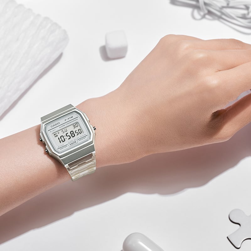 La montre-bracelet F-91WS-7 argentée et transparente sur une table à côté d'objets blancs/transparents tels qu'un câble de recharge et une pièce de puzzle.