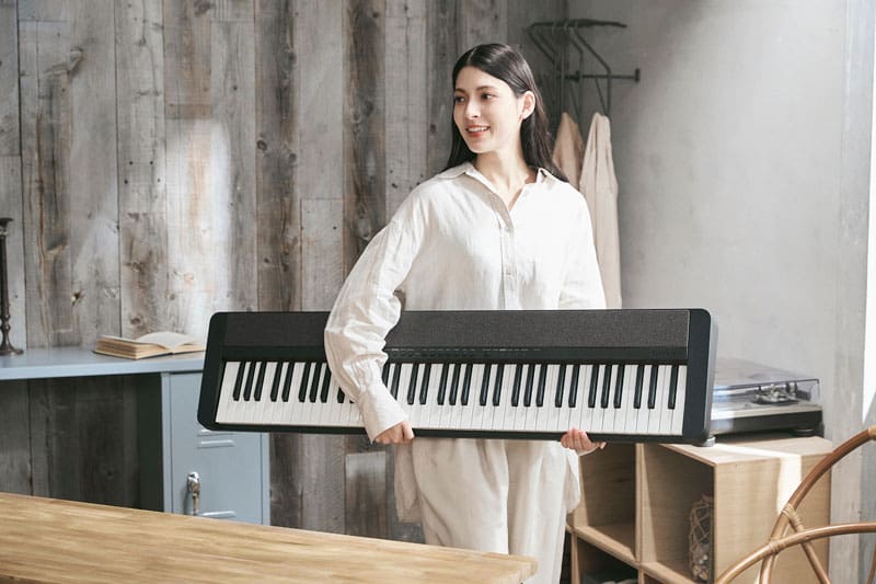 Une femme vêtue de couleurs claires porte un piano numérique CT-S1-76BK noir sous un bras dans un bureau confortable