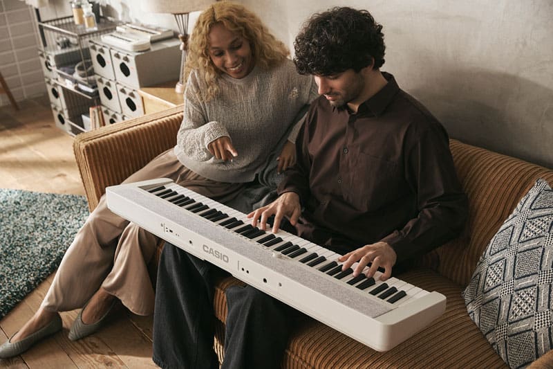 Une jeune femme et un jeune homme sont assis sur un canapé marron. Un piano numérique CT-S1-76WE blanc est posé sur les genoux de l'homme. Il joue du piano et la femme, souriante, commente le jeu d'un geste du doigt.
