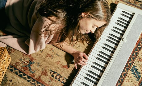 Une jeune femme est allongée sur un tapis et joue sur le piano numérique blanc CT-S1WE qui est posé sur le sol devant elle.