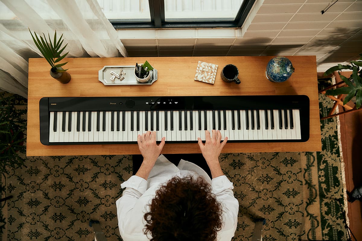 Vue d'en haut du piano numérique Privia PX-S5000, posé sur un étroit bureau en bois. Une personne est assise devant et joue du piano.