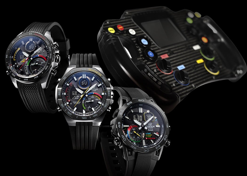 Tre orologi da polso neri i cui quadranti sono caratterizzati dai quattro colori d'accento rosso, verde, blu e giallo; sullo sfondo si intravede il volante di un'auto da corsa con pulsanti colorati
