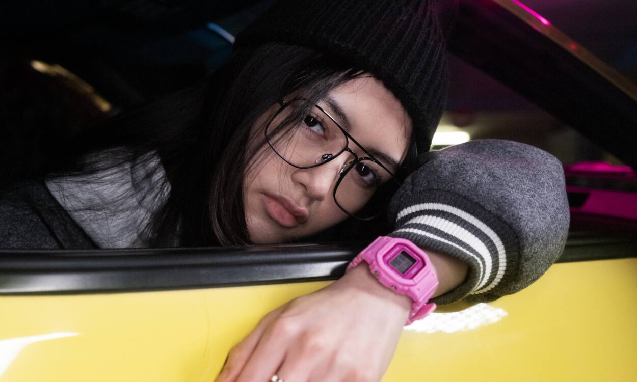 Una donna che scende da un'auto gialla dal sedile del passeggero, con un cappello nero, occhiali e l'orologio digitale GMD-S5600PP rosa di G-SHOCK.