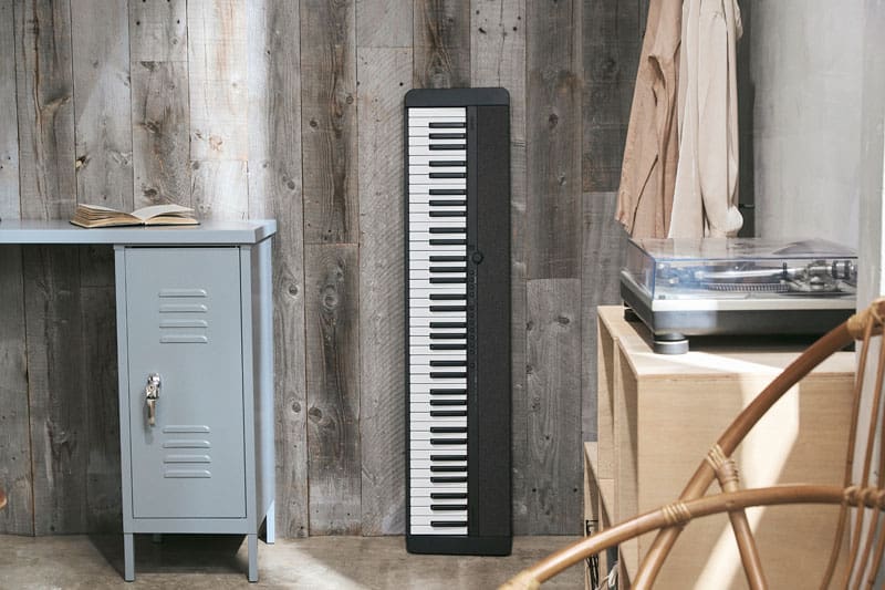 Un pianoforte digitale nero CT-S1-76BK è appoggiato in posizione verticale su una parete grigia di assi di legno, accanto a una scrivania azzurra
