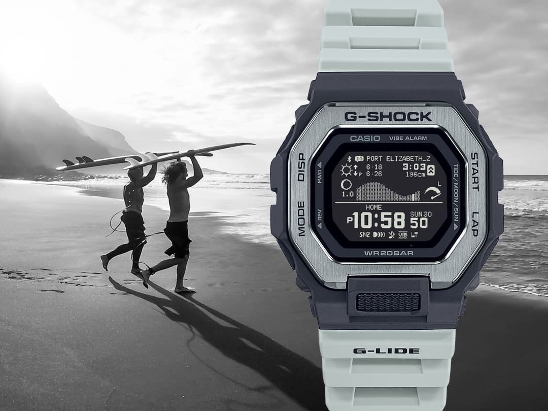 Voorkant: het Casio G-Shock GBX-100TT-8 polshorloge met lichtgrijs kunststof bandje, zwarte kast en zilverkleurige lunette, achterkant: een zwart-wit afbeelding van twee surfers die over het strand de zee in rennen