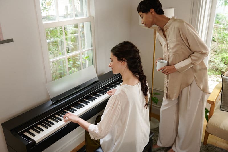 Een vrouw bespeelt een zwarte AP-S200BK digitale piano terwijl een tweede vrouw achter haar staat en toekijkt hoe ze speelt