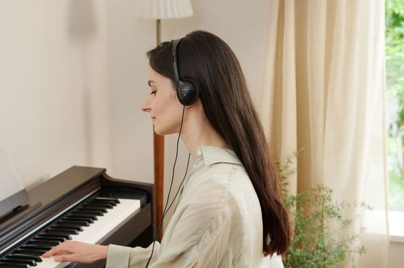 Een vrouw met een koptelefoon op zit glimlachend bij een donkere AP-S200 digitale piano en speelt piano