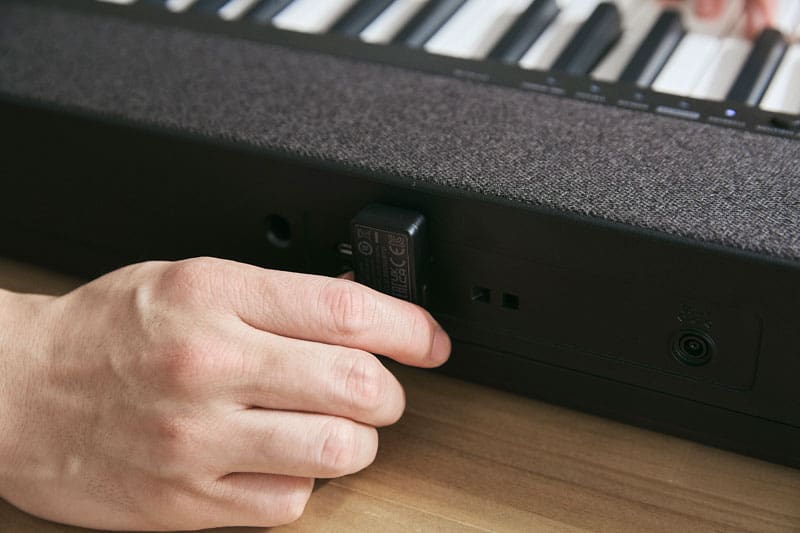Close-up van de achterkant van de zwarte CT-S1-76BK digitale piano met verschillende aansluitingen en een hand die een adapter insteekt