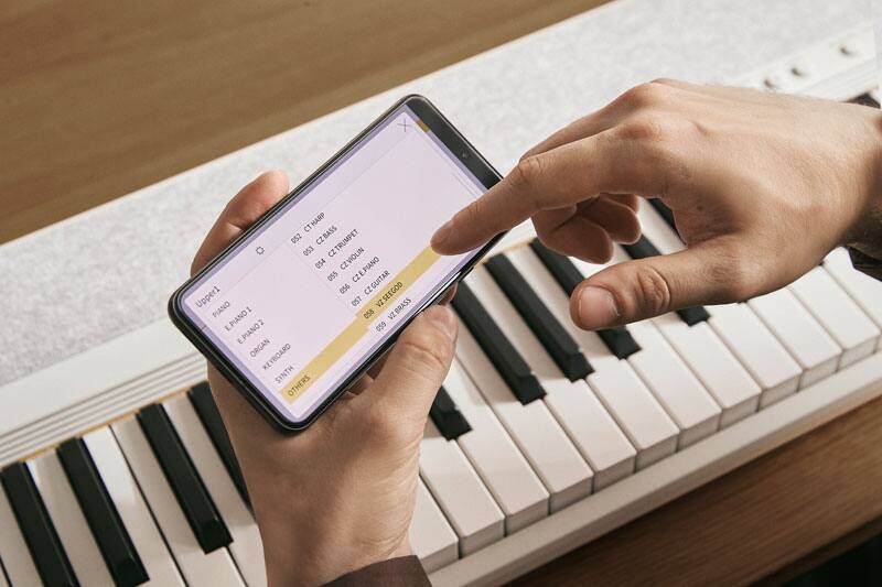 Bovenaanzicht van een witte CT-S1-76 digitale piano en een hand die een smartphone vasthoudt terwijl de andere hand de geluidsinstellingen aanpast in de CASIO MUSIC SPACE app