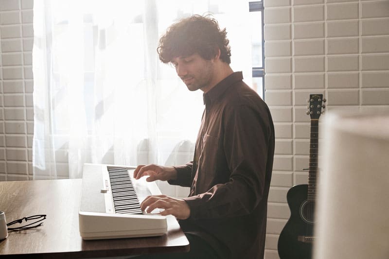 Een man in een donker overhemd zit tegen het licht in aan een tafel voor een raam en bespeelt de witte CT-S1-76WE digitale piano
