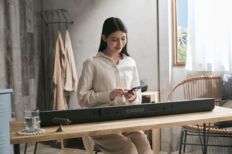 Een jonge vrouw in een lichtgekleurde, eenvoudige blouse zit aan een tafel voor een CT-S1-76BK digitale piano. Ze kijkt op haar smartphone, die ze in haar linkerhand houdt