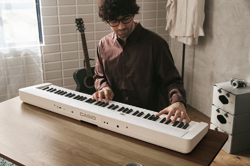 Een man in een bruin overhemd aan een bruine tafel bespeelt de witte CT-S1-76WE digitale piano