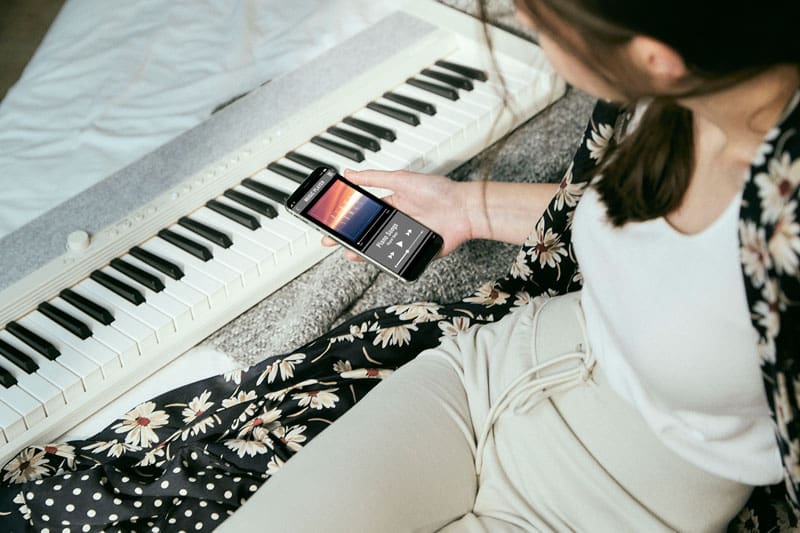 Een vrouw zit naast een witte CT-S1WE digitale piano en kijkt naar haar smartphone