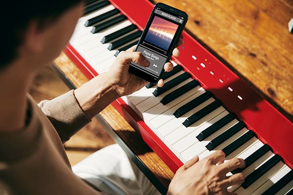 Een persoon speelt met zijn rechterhand op de rode Privia PX-S1100RD digitale piano terwijl hij naar zijn smartphone kijkt, die hij in zijn linkerhand houdt