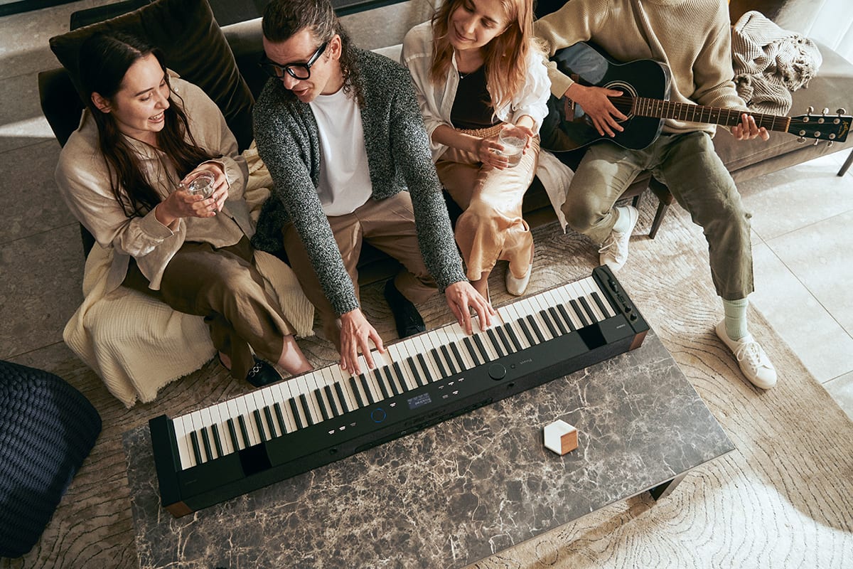 Vier jonge mensen zitten op een sofa, een van hen speelt op de PX-S6000 digitale piano voor hem op een woonkamertafel, een ander speelt gitaar