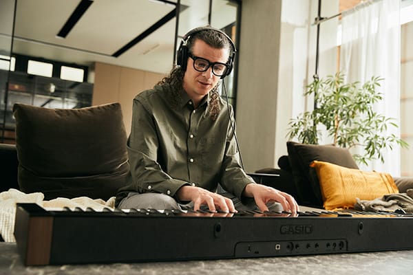 Gezicht van voren op een jonge man die een koptelefoon draagt en piano speelt op de digitale piano PX-S6000 die voor hem op de tafel in de woonkamer ligt
