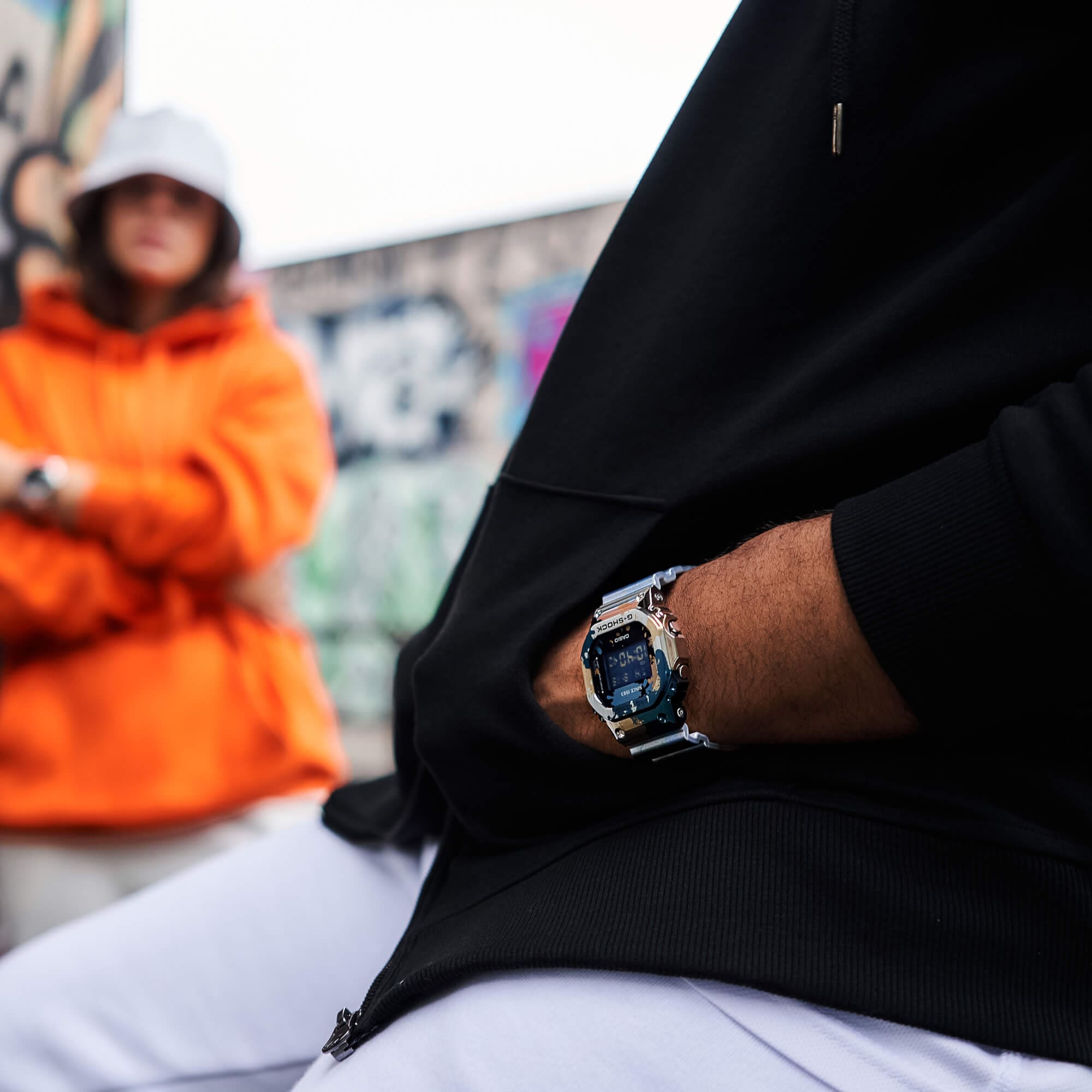 Op de voorgrond draagt een persoon een zwarte trui met aan zijn pols het GM-5600SS-1ER horloge van G-SHOCK.