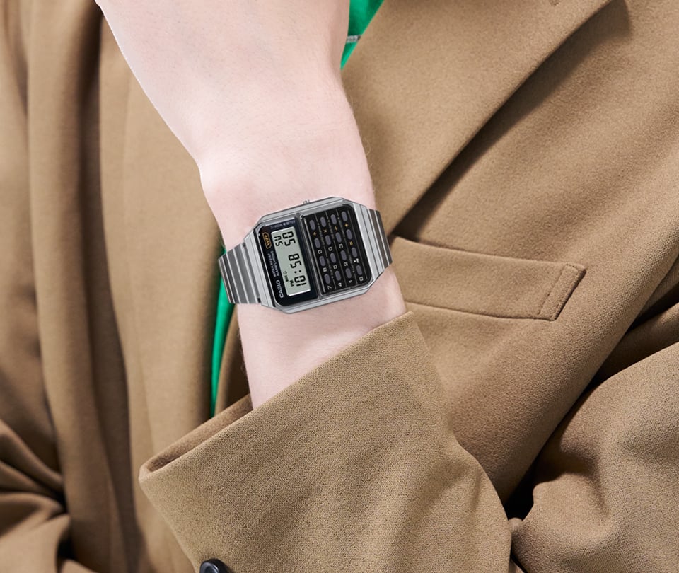 CA-500WE-1A O relógio em aço inoxidável prateado CA-500WE-1A da coleção Casio Vintage no pulso de uma pessoa com um blazer cor de caramelo sobre uma t-shirt verde