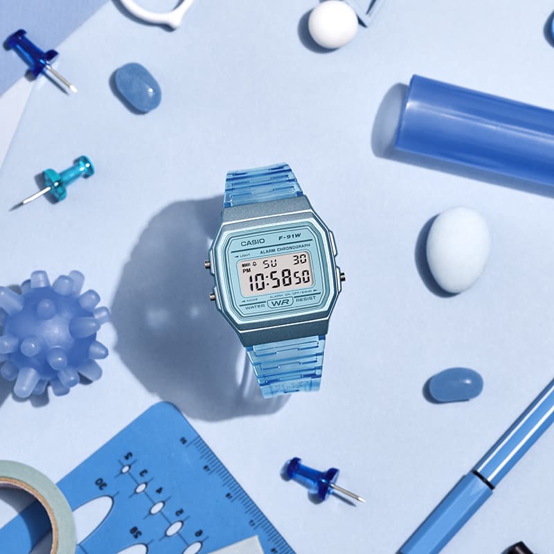 O relógio de pulso azul transparente F-91WS-2 sobre uma mesa ao lado de objetos azuis, como um alfinete e uma caneta.