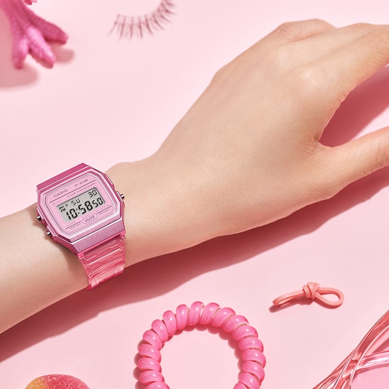 O relógio de pulso F-91WS-4 rosa transparente sobre uma mesa ao lado de objetos rosa, como uma fita para tranças ou óculos de sol.