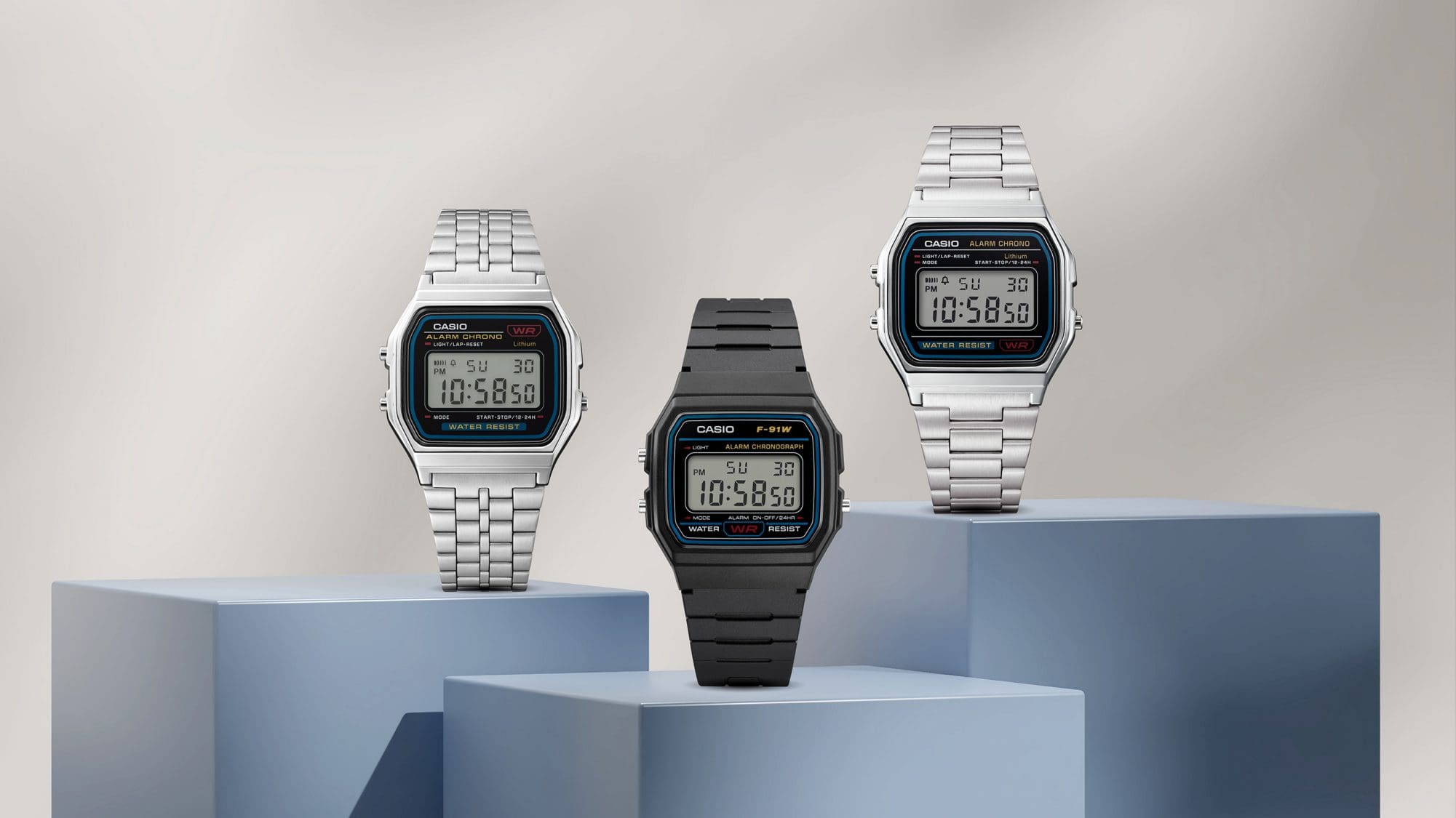 Três modelos diferentes do F-91W da CASIO, com bracelete prateada ou preta em pedestais.