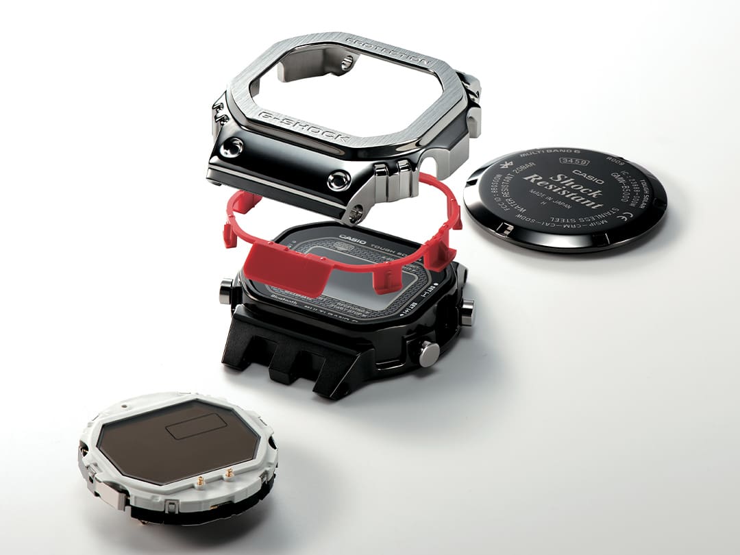  A luneta prateada em aço inoxidável, o componente amortecedor vermelho e a caixa preta são mostrados um em cima do outro, com a parte de trás da caixa e o ecrã ao lado