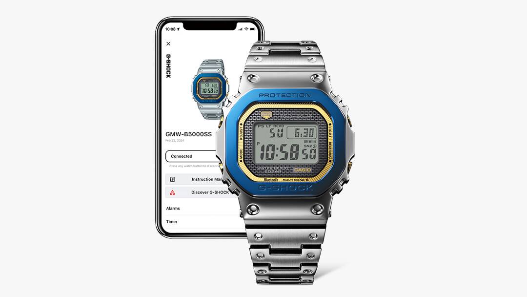 O relógio de pulso de aniversário Casio G-Shock GMW-B5000SS-2 com bracelete prateada em aço inoxidável, luneta azul e mostrador dourado em frente a um smartphone com informações sobre o relógio no ecrã