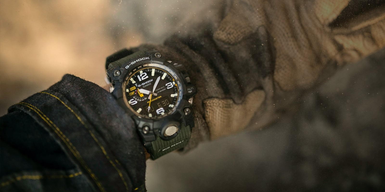 Um relógio de exterior Master of G Mudmaster GWG-1000-1A3 num pulso entre as mangas de um casaco de ganga e luvas de trabalho