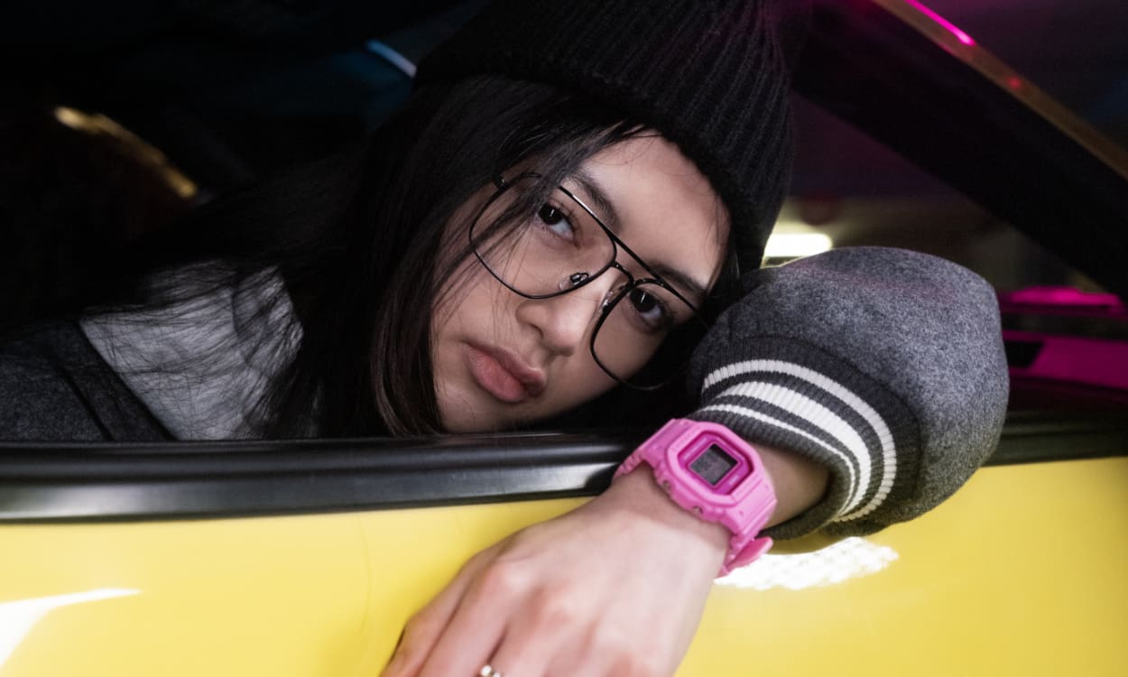 Uma mulher inclina-se para fora de um automóvel amarelo a partir do banco do passageiro, com um chapéu preto, óculos e o relógio digital GMD-S5600PP cor-de-rosa da G-SHOCK.