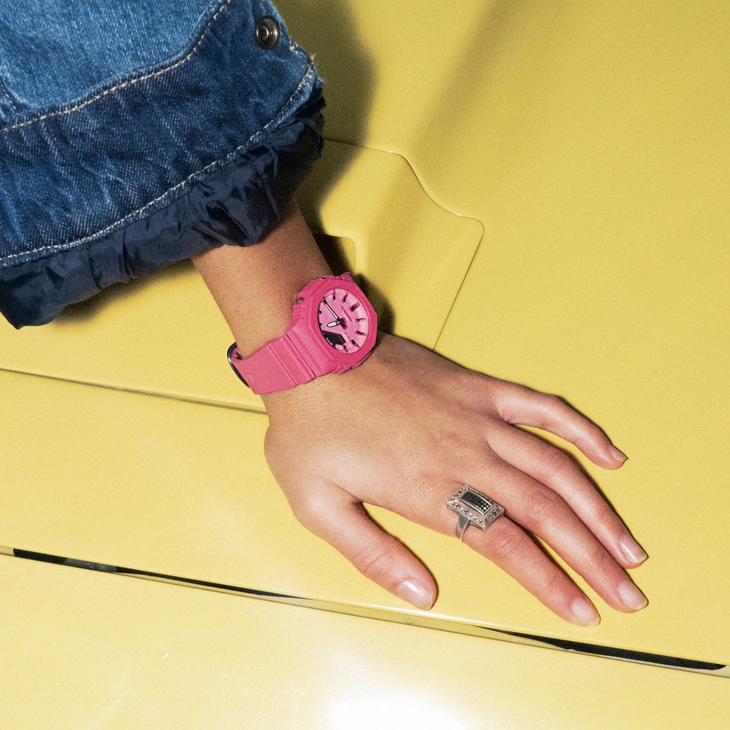 A mão de uma mulher apoiada no capô de um motor amarelo, usando o relógio analógico cor-de-rosa GMA-2100PP-4