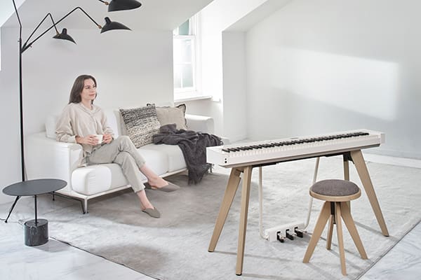 Uma mulher senta-se num sofá de uma sala de estar bem iluminada e olha para o piano digital PX-S7000WE branco
