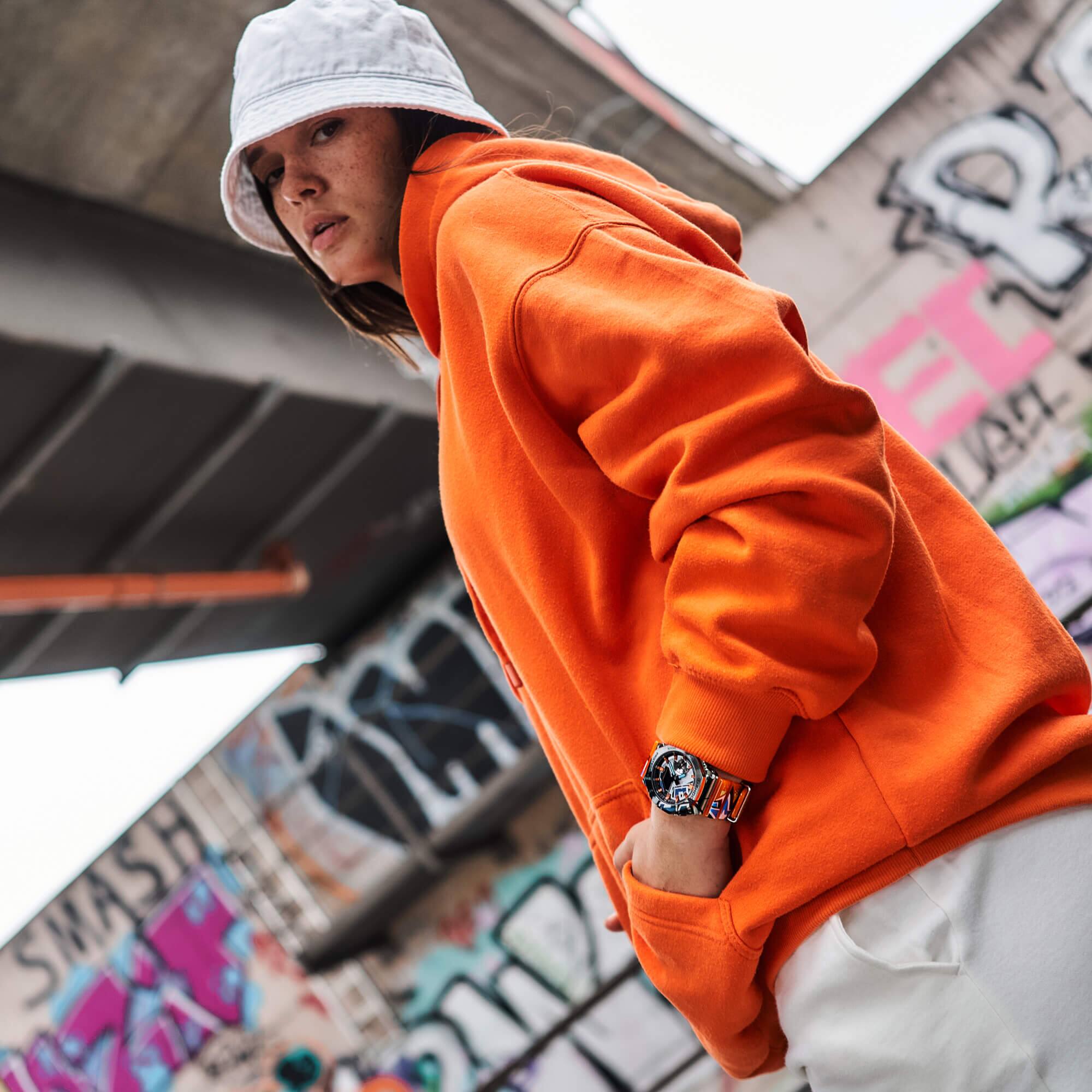 Da perspectiva do sapo: Uma mulher com uma cabeça de balde branca usa um capuz laranja e o GM-2100SS com bracelete laranja da G-SHOCK.